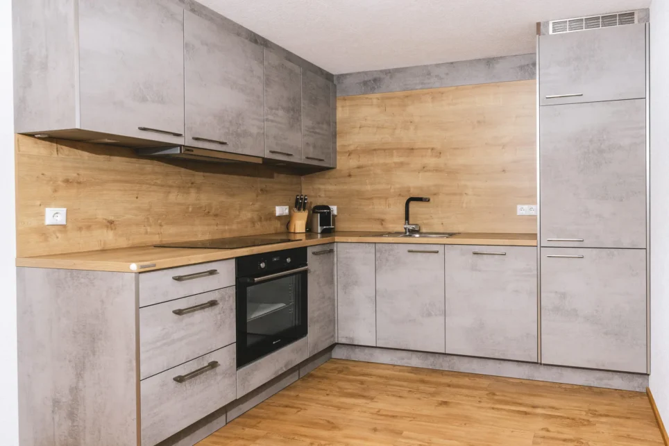 Eckküche eines Appartements: graue Fronten, Holzrückwand und Arbeitsplatte, Einbauherd, Spüle, Kaffeemaschine, Holzboden